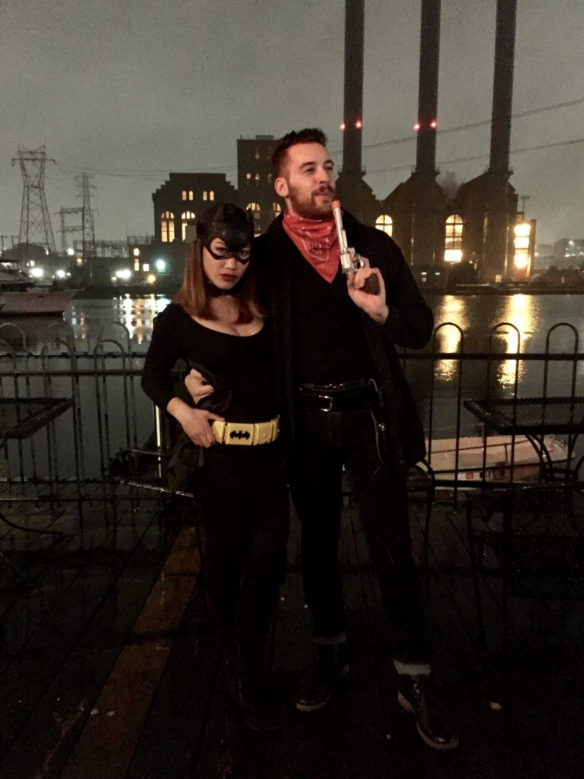 Josh and Sophia posing in costume. Josh is dressed as a cowboy and Sophia's Batwoman. A scenic river with Industrial buildings can be seen in the background.